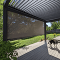 A black Bon Pergola, with motorized louvered roof panels, stands over a patio decked in light gray wood. One side of the pergola features a dark-colored sunshade screen. The patio is open and uncluttered, with just a small dog wandering through the sunlight.