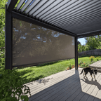A black Bon Pergola, with motorized louvered roof panels, stands over a patio decked in light gray wood. One side of the pergola features a dark-colored sunshade screen. The patio is open and uncluttered, with just a small dog wandering through the sunlight.