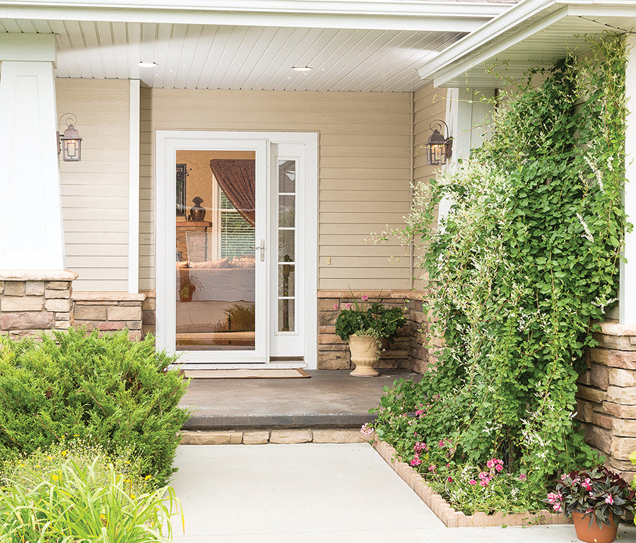 A white full view aluminum storm door installed on a home entrance, with surrounding greenery and a potted plant.